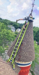 A man is grabbing onto the apex of a cone shaped roof from the near top of a bright yellow ladder. He has on a safety harness and ropes.