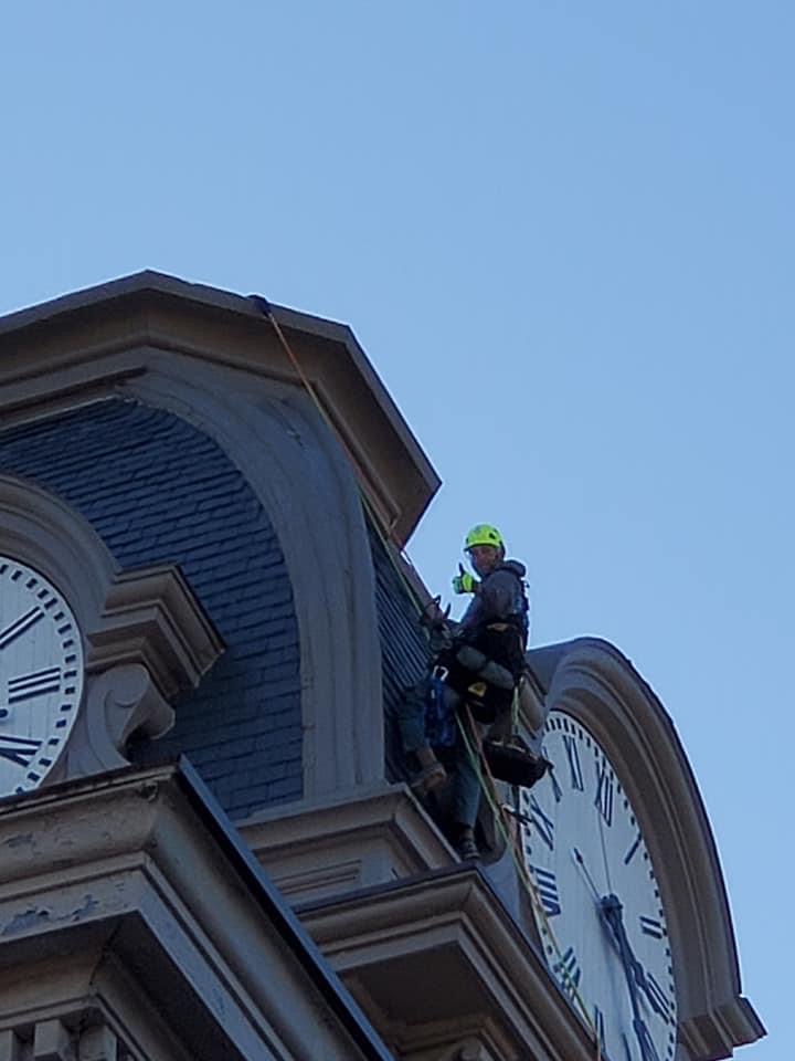 A man attached by harness and ropes from the top of a clocktower. The man has on bright yellow safety gear and a helmet, and is giving a thumbs up to the camera.