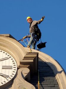 A man standing on the top of a clocktower. He is attached to and holding onto the building by rope and harness. Other safety gear is visible on him. He's waving to the camera.