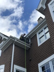 A man is sat between two levels of a joining roof, between gutters. There is a safety rope visible.