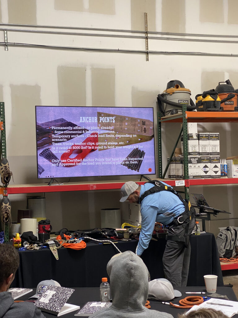 A man wearing a safety hardness is standing in front of a group of seated students. He has a table full of ropes and various safety equipment. There's a tv displaying a slide show behind him, displaying "ANCHOR POINTS" and associated images and text.