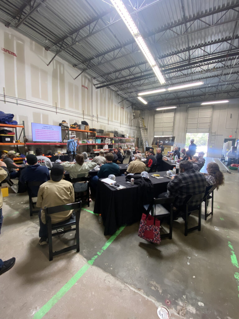 Wide angle shot of a warehouse. There are around 30 people seated at tables with notebooks and branded hats. In front of them, a man in a safety harness is holding a rope.