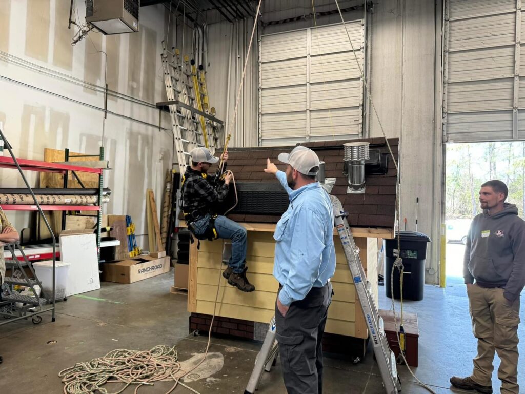 A man wearing a safety harness is suspended from the out of shot warehouse roof. He's next to a scale building with roof. An instructor next to him is pointing.