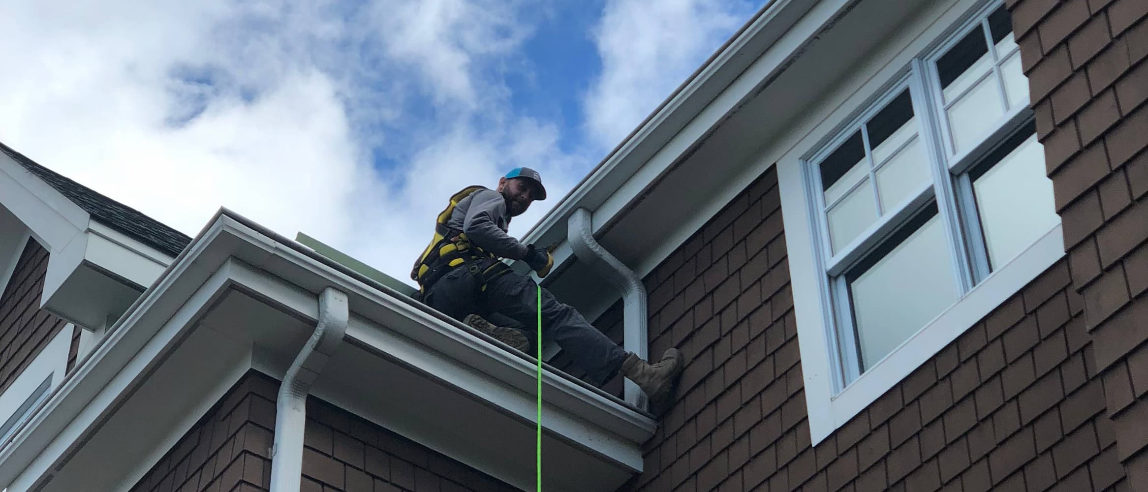 A man is sat between two levels of joining roof, between two gutters facing different directions. He is wearing a safety harness and other equipment. It looks like he's giving the camera a thumbs up.