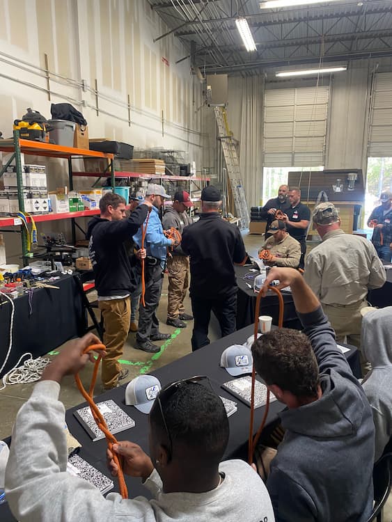 A group of people are standing in a warehouse working with ropes, in the process of tying knots in them. Some people are sitting near the camera doing the same, with branded hats and notebooks on their table.