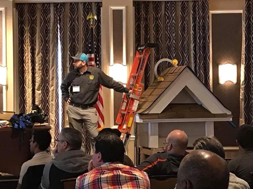 A man is in front of a class of at least 8 other people. He's holding onto a small ladder which is leaning against a scale house. They're all looking off camera.