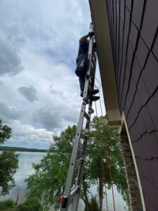 A person near the top of a ladder, which is leaning against a house overlooking a lake. There is visible safety equipment on both the ladder and the person's body.