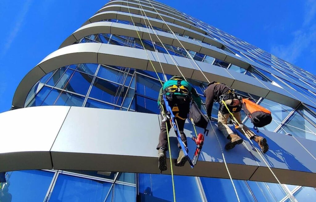 Two people are attached by ropes and harnesses on the side of a tall glass sided building. They have safety gear and tools attached to them.