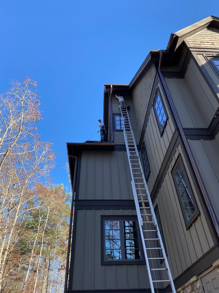 A tall ladder leaning against the side of a 4 story building. A man is near the top of the ladder and another is standing on the 3rd story roof next to him. Safety equipment is visible on both men.